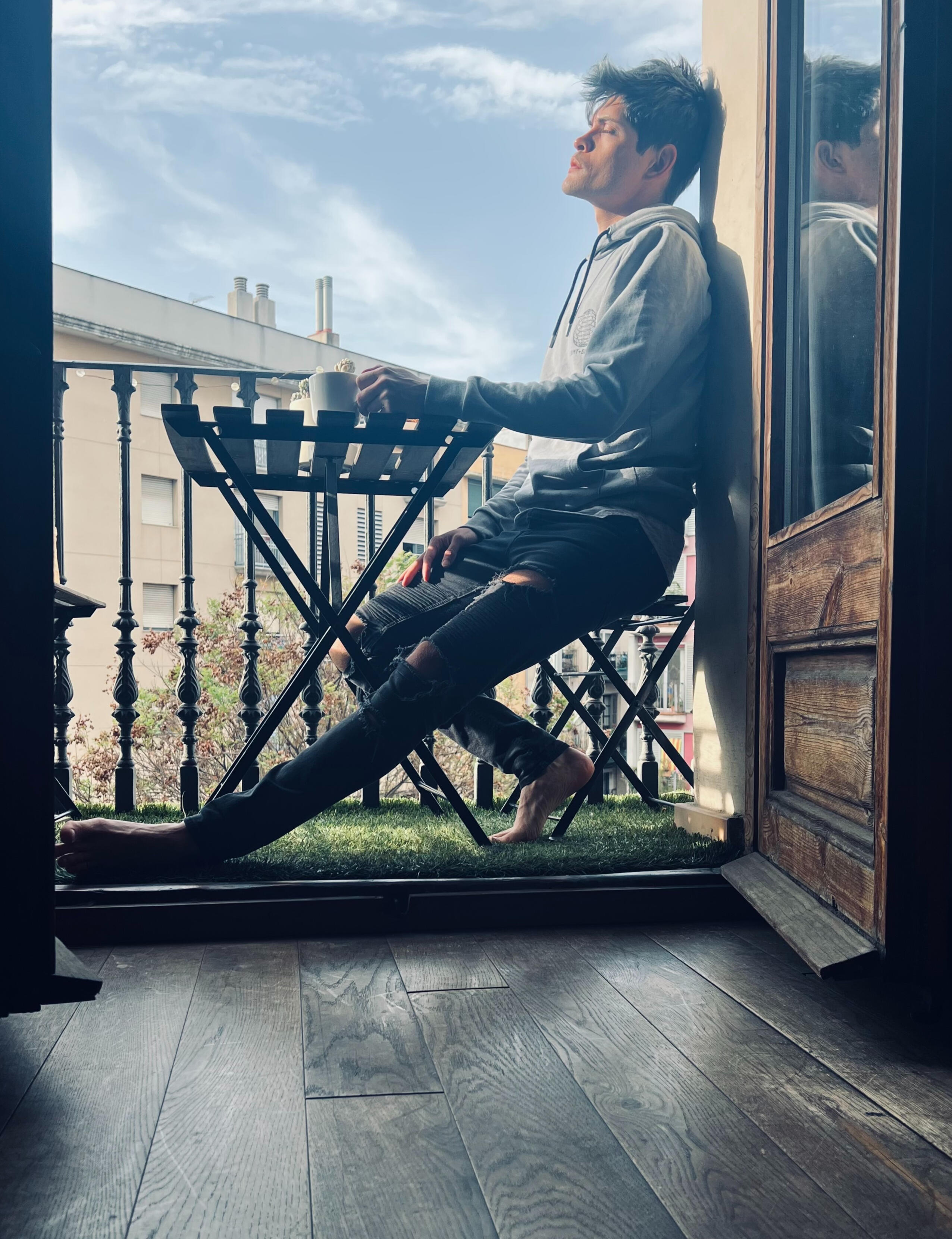 A very tired and overstimulated human is wearing yesterday's grey hoody and ripped black skinny jeans. They're enjoying the morning sun with eyes closed against a door on a balcony.