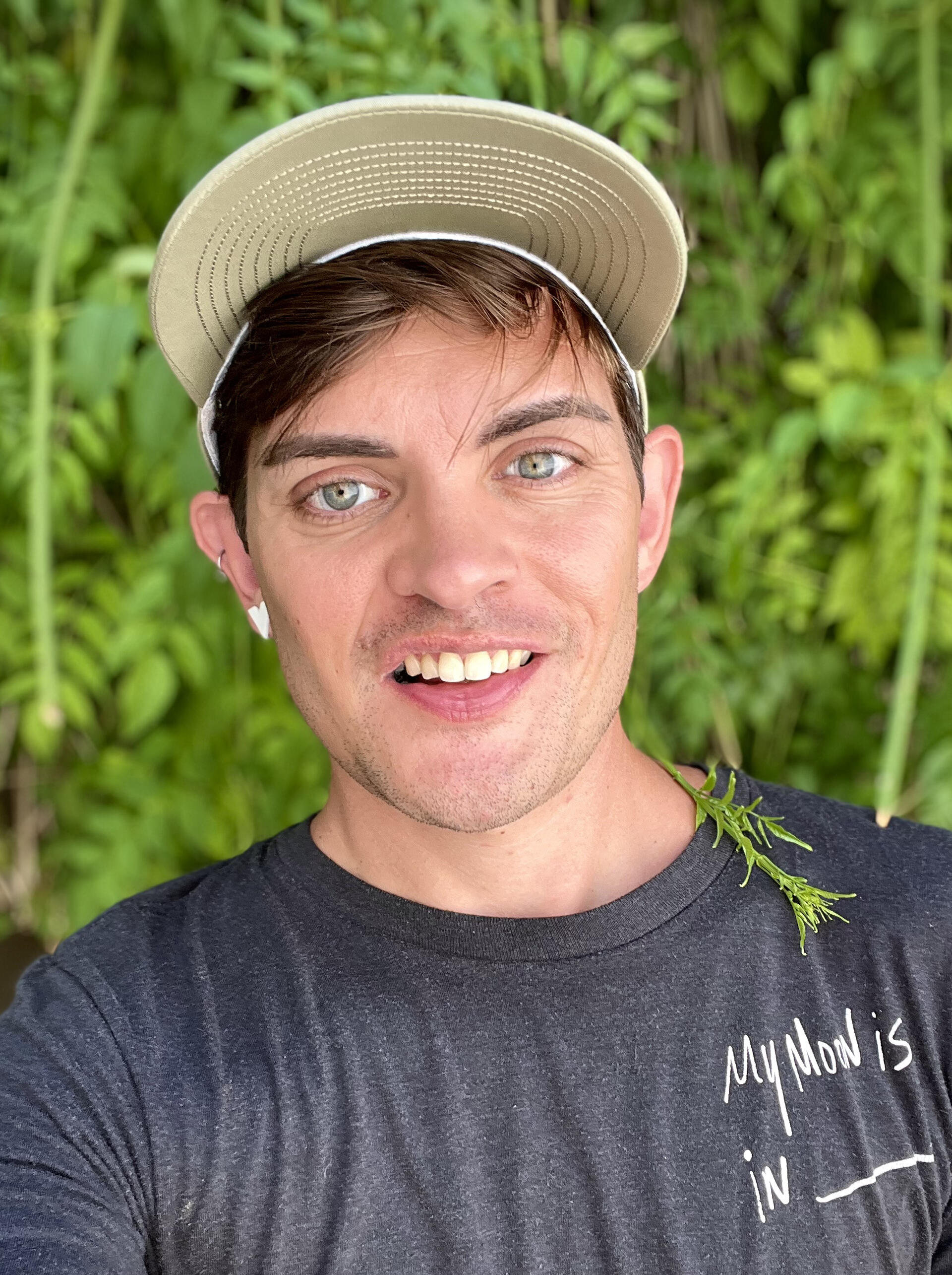 A cutie patootie human in a beige cap and gray shirt smiles at the camera against a lush green background of plants.